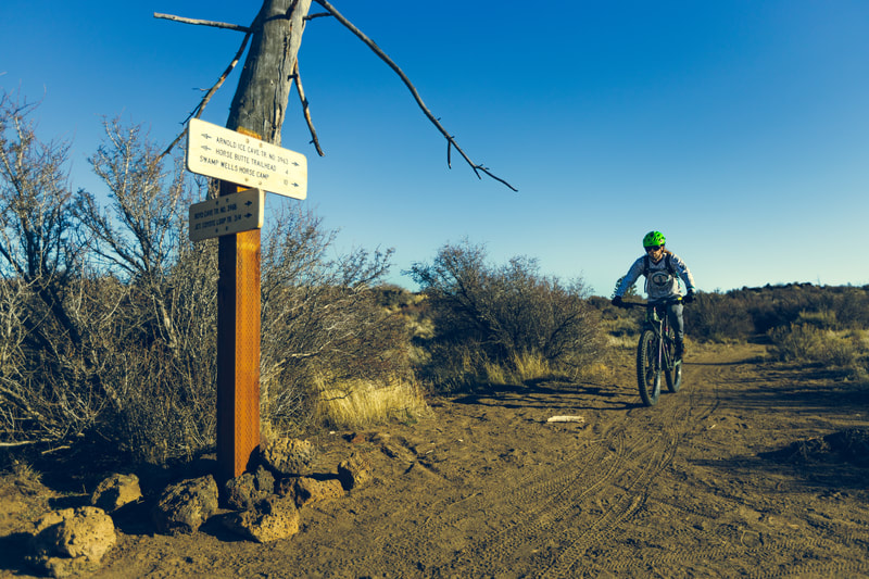 Biker riding up to an intersection with Arnold Ice Cave, Horse Butte trailhead, Swamp Wells trails on one sign, and Boyd Cave and Coyote Loop trails on the other